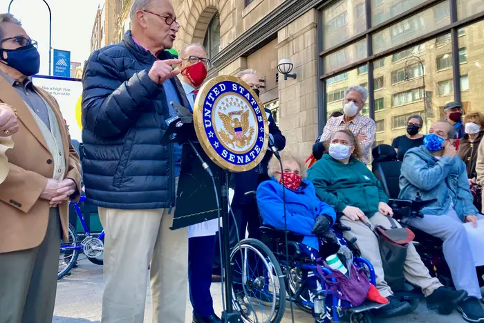 Senator Chuck Schumer, wearing a mask, speaks at a podium outside the 14th Street and 6th Avenue subway station to urge the federal government to release funding to build 6 elevators at the station.
